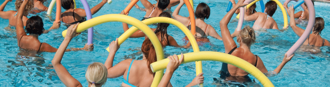 A group of ladies doing aquatic excercise with pool noodles raised above their heads.